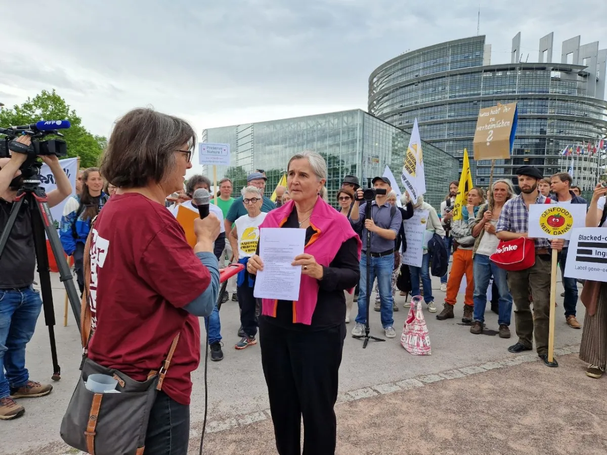 Protest vor dem Gebäude des Europäischen Parlaments in Straßburg - im Hintergrund Aktive mit Transparenten, im Vordergrund ein Interview (eine Frau mit Mikro, eine Frau mit Flugblatt) und Pressevertreter*innen mit Kameras